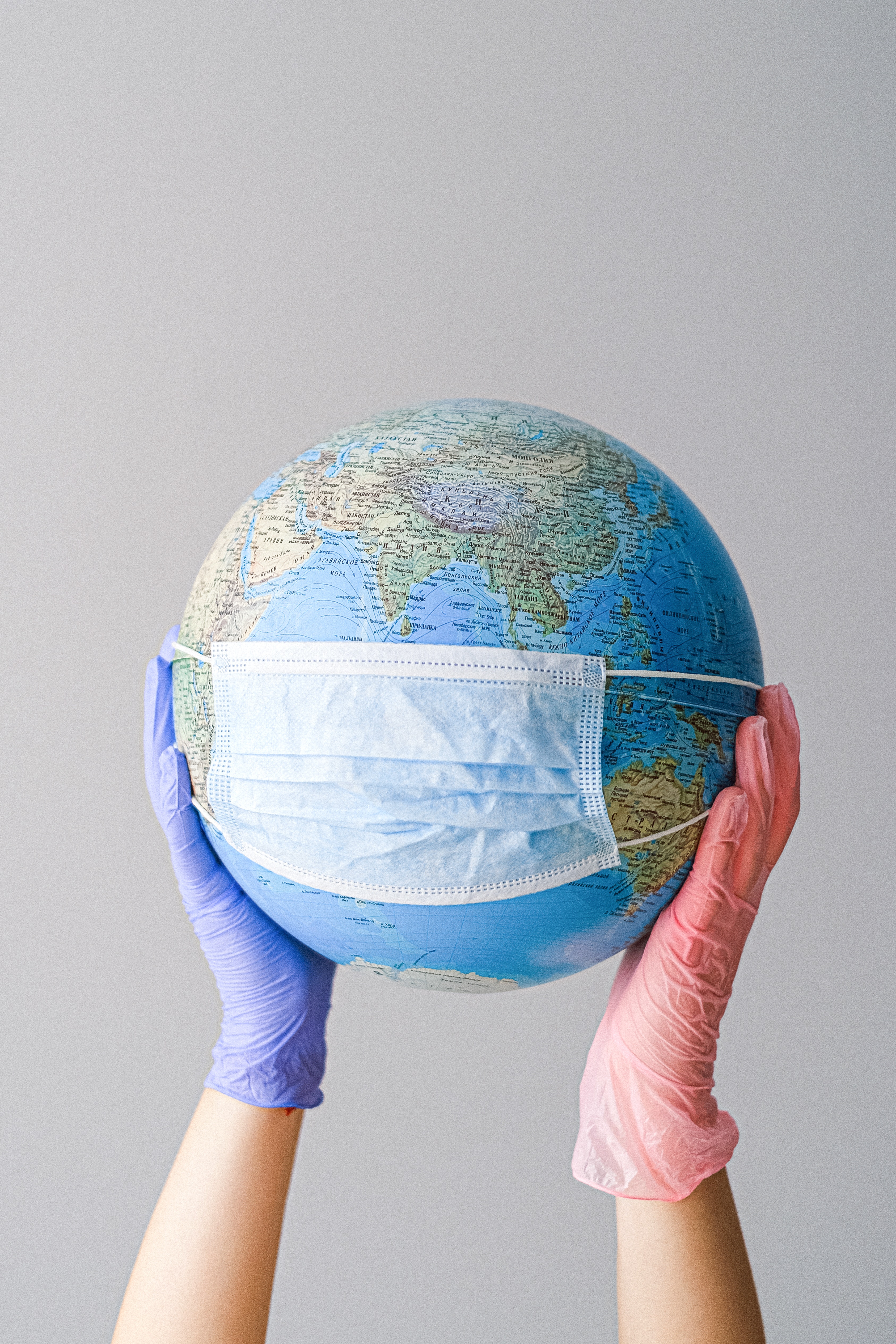 A globe wearing a mask, held up by 2 hands, one wearing a blue glove and the other wearing a pink glove.
