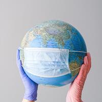 A globe wearing a mask, held up by 2 hands, one wearing a blue glove and the other wearing a pink glove.